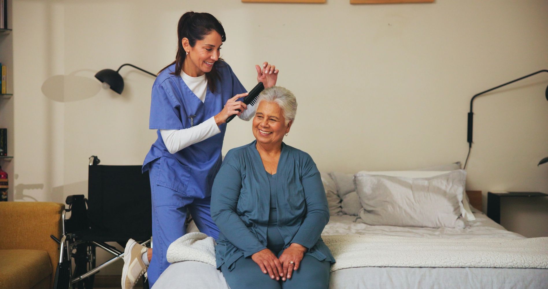 A smiling caregiver in blue scrubs gently combs the hair of an older woman.