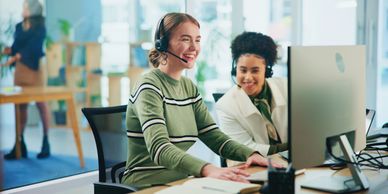 Two women with headsets smiling at a computer in a bright office.