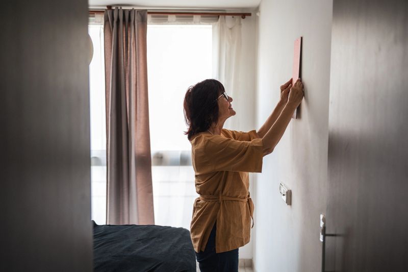 A woman decorates her bedroom by hanging artwork on the wall, creating a personalized and cozy atmosphere. The bedroom features soft natural light and a modern aesthetic.