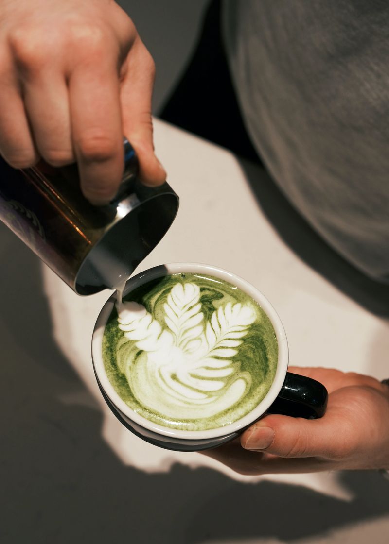 A close-up shot of a barista pouring steamed milk into a cup of vibrant green matcha latte, forming delicate latte art. The rich, earthy color of matcha blends beautifully with the creamy milk, creating a visually appealing drink. This popular beverage is enjoyed in cafes worldwide for its health benefits and smooth taste. Ideal for coffee shop branding, menu designs, and beverage lifestyle photography.