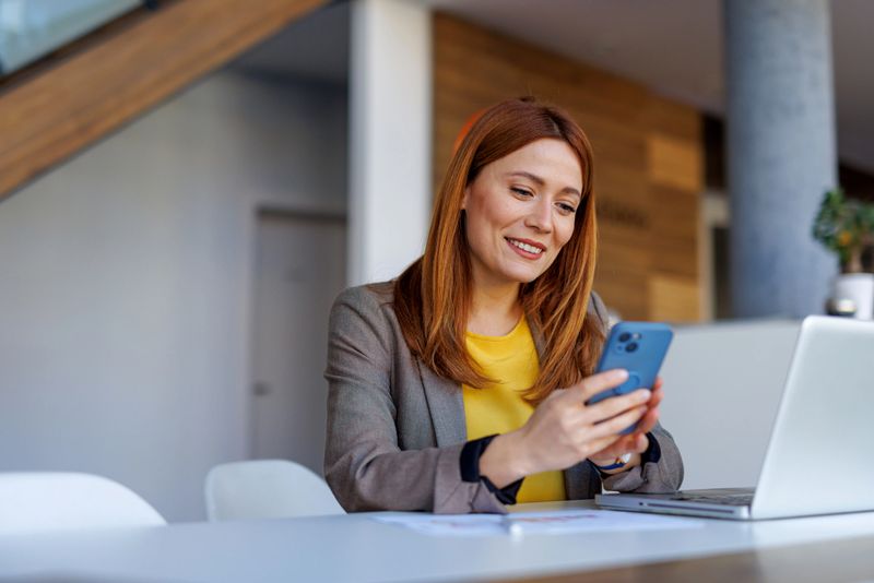 Businesswoman sitting at a modern desk in a bright office, smiling while using a smartphone and working on a laptop, showcasing productivity and professional success