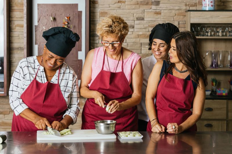 Four cheerful women wearing aprons preparing ingredients for a recipe, having fun together during a cooking course