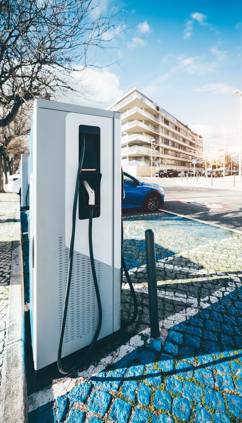 Electric vehicle charging station with a black charging cable and plug attached to a modern white charger unit in an urban setting. A blue car is parked nearby, with residential buildings behind