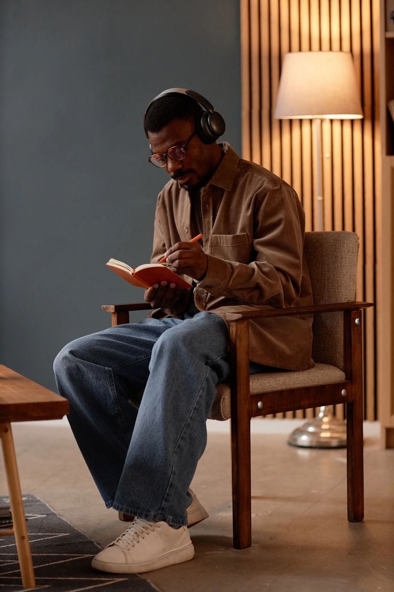 Vertical full length of Black man dressed in casual outfit immersed in writing work notes, while listening to music through headphones sitting in vintage armchair in home office