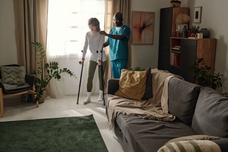 Healthcare professional assisting patient in standing exercise in comfortable living room setting with natural light and indoor plants enhancing serene atmosphere