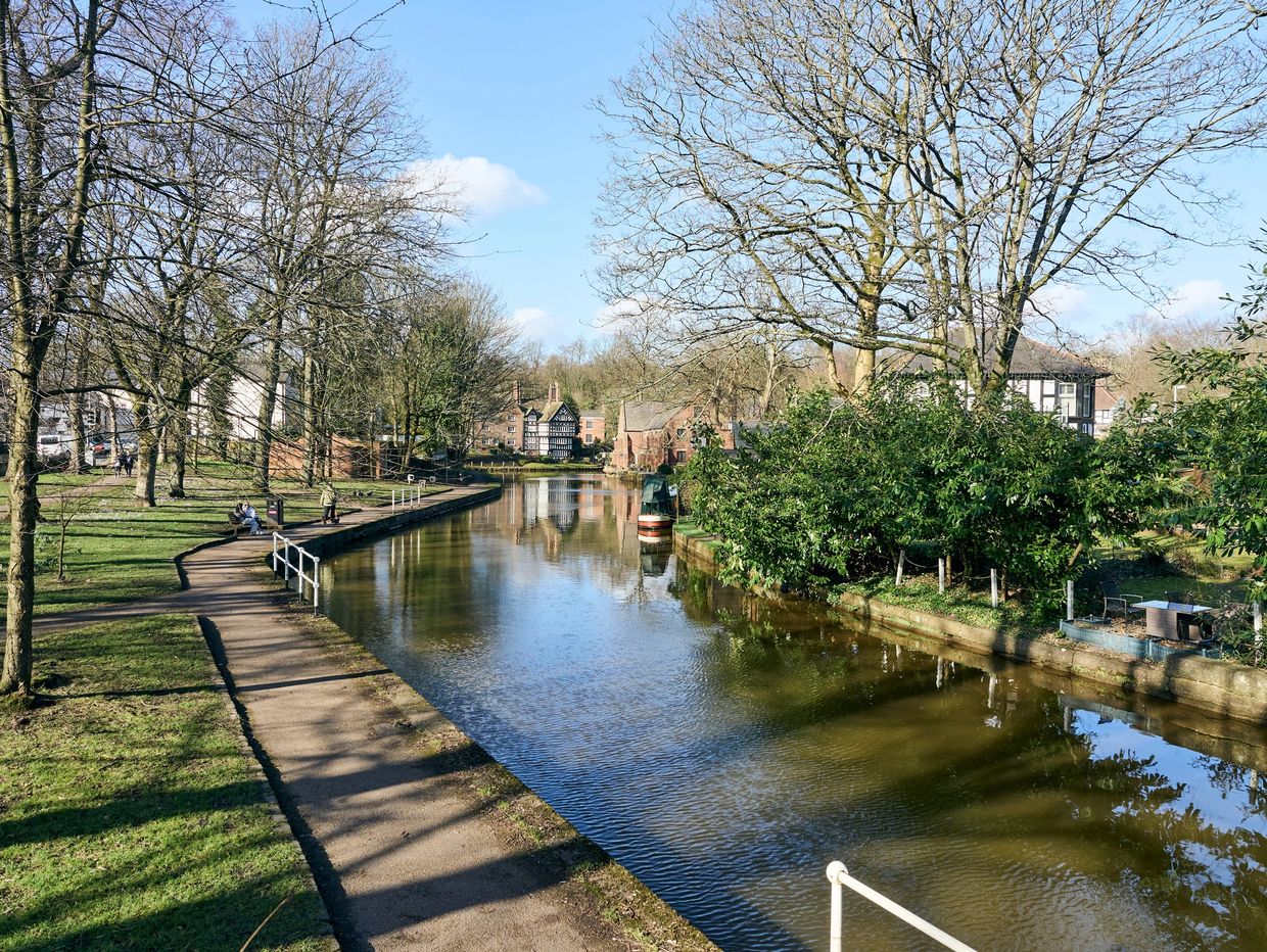 A peaceful riverside path with trees and historic houses under a clear blue sky.