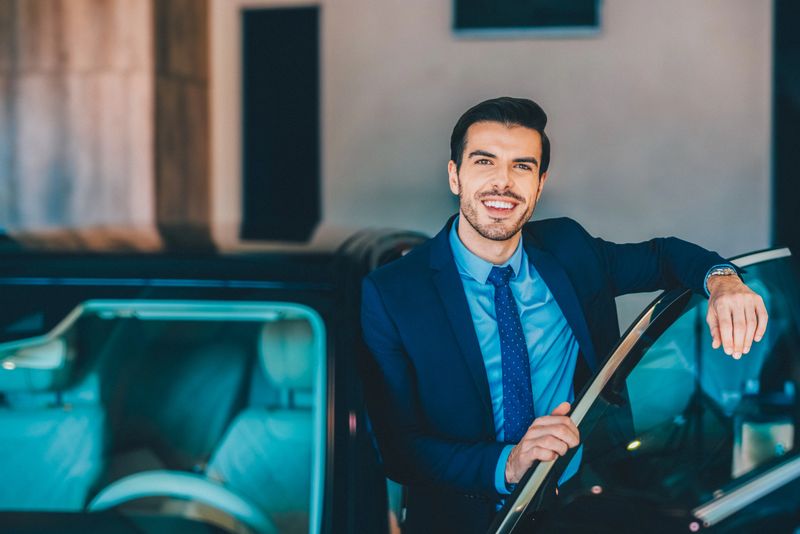 Businessman in a navy suit and tie confidently leans on the door of a luxury car, exuding professionalism and success.