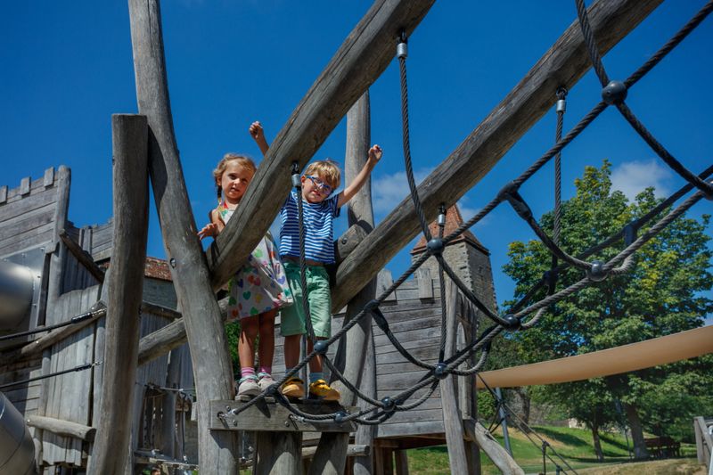 Two young children are enjoying a wooden play structure, with the sun shining brightly against a cloudless sky