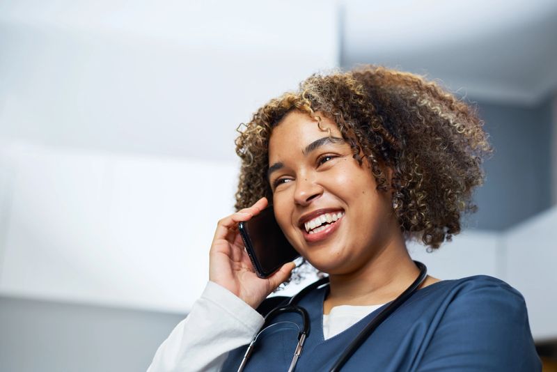 A cheerful healthcare worker, wearing medical scrubs and a stethoscope, chats on a phone call in a bright clinic environment. The image conveys professionalism, approachability, and effective communication during healthcare services.