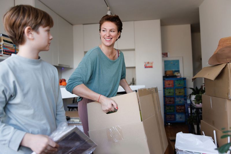 Cheerful mid adult woman carrying cardboard box, her teenage son helping and bringing items for packing, family moving house