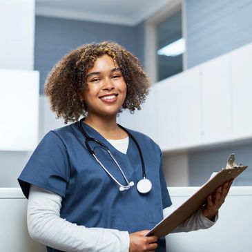 Smiling nurse in blue scrubs holding a clipboard in a medical setting.