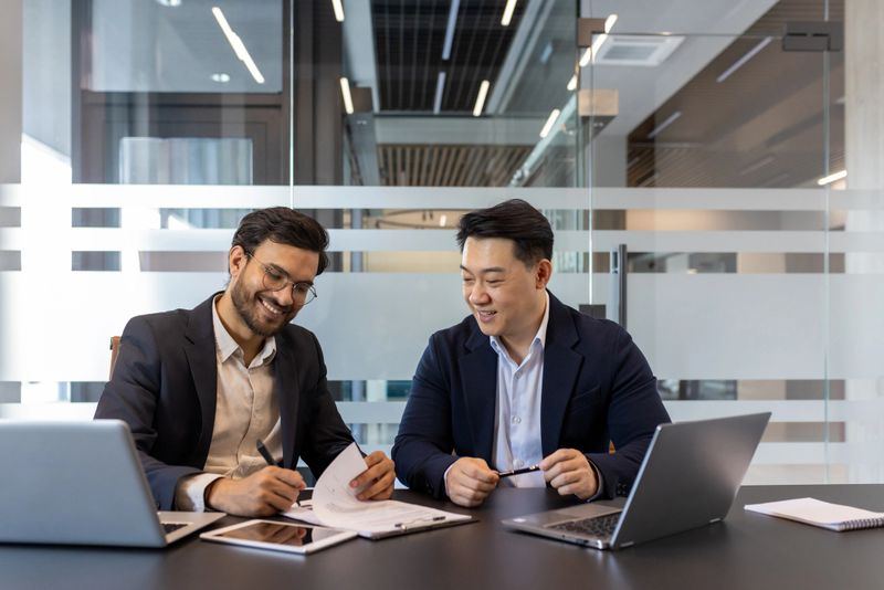 Two professionals are working together, reviewing documents in a modern conference room setting. They are surrounded by laptops and other tools, engaging in a discussion.