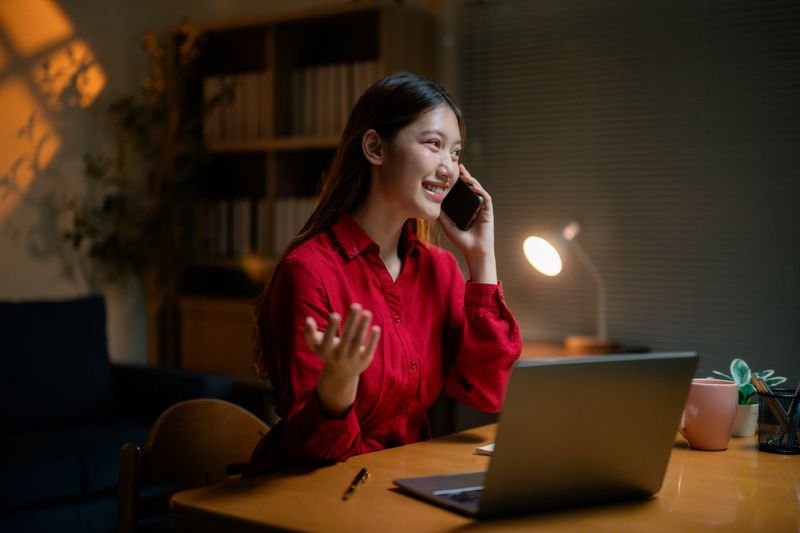 Young Asian businesswoman wearing red shirt talking on smartphone and working late at night in home office using laptop computer, desk lamp illuminating the space