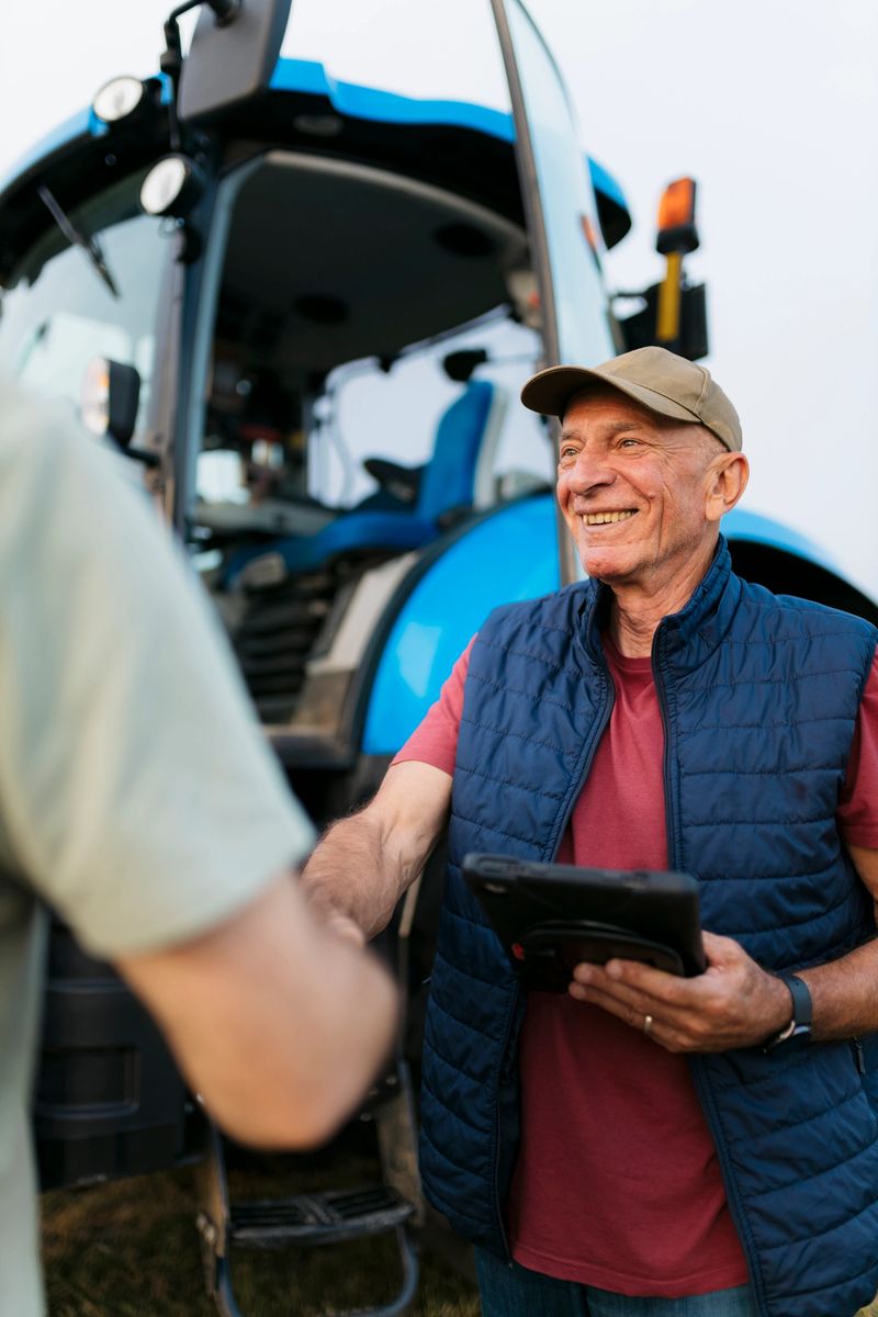 Smiling senior farmer holding a digital tablet and shaking hands with a man beside a tractor on an agricultural farm. Happy elderly man meeting a farm worker on farm.