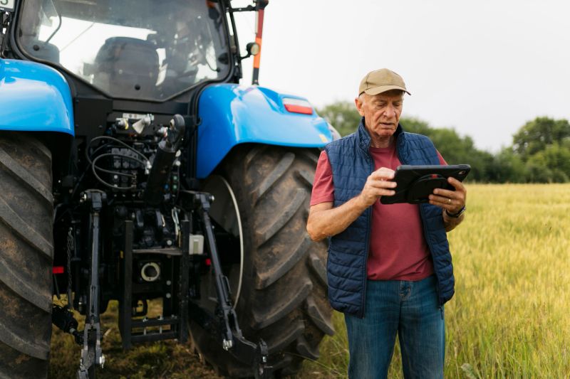 Senior farmer utilizing a digital tablet on his farmland with his tractor in the background, illustrating the integration of digital tools in agriculture.