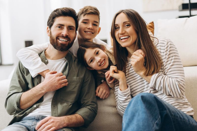 Cheerful family of four, including a father, mother, son, and daughter, is sitting together on a comfortable sofa in their living room, embracing each other and smiling happily