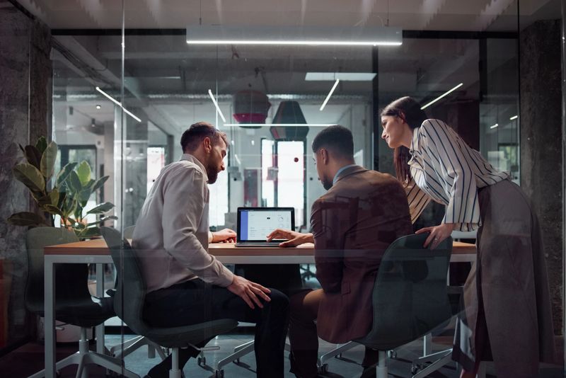 A group of coworkers discuss ideas at a laptop in a modern glass-walled workspace, emphasizing teamwork and professional collaboration. The office fosters creativity and productivity with its open and interactive environment.
