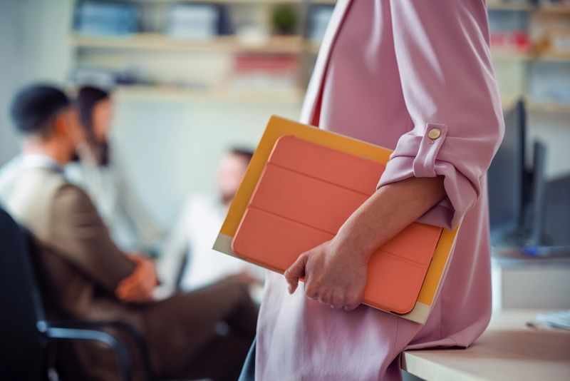 Close-up of a professional woman holding files while colleagues in the office discuss business projects in the background, highlighting teamwork and organization in a corporate environment.