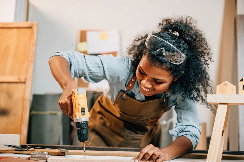 Carpenter woman using a power drill to secure a piece of wood in a workshop, focusing on precision in furniture making. Surrounded by tools and wood pieces, highlighting skill and craftsmanship