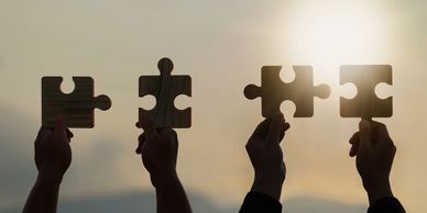 Four hands hold wooden puzzle pieces against a sunset sky.