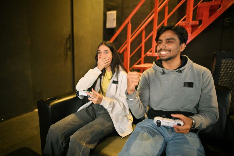 A young man celebrates his video game victory with excitement while his female friend sits beside him with a shocked expression in a lively gaming café. Both are holding controllers, immersed in the game, surrounded by glowing screens and neon lights. The contrast between the winner’s joy and the loser’s frustration captures the competitive spirit and fun dynamics of gaming with friends in an energetic gaming hub.