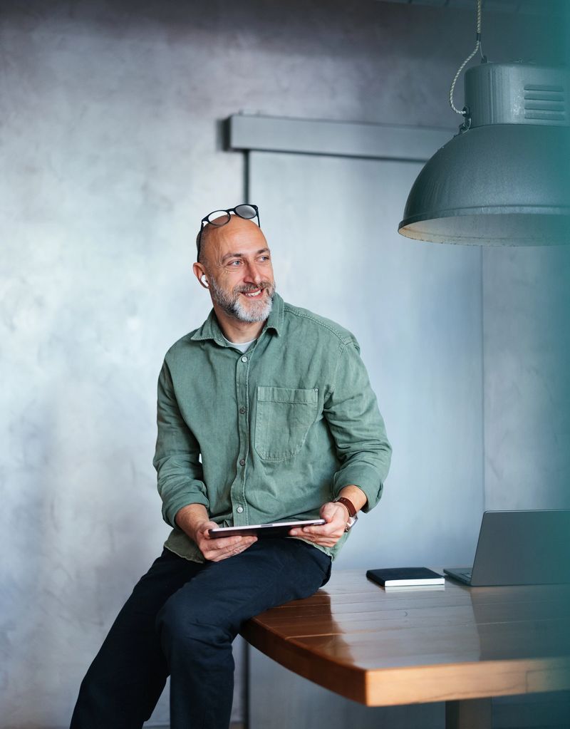 Bald man in a green shirt leaning on a desk, working with a digital tablet and laptop inside a modern workspace showcasing a relaxed and productive atmosphere.