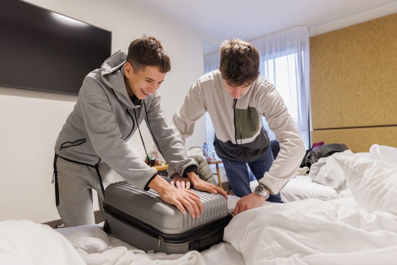 Two teenage boys having fun and closing packed suitcase on the bed before traveling