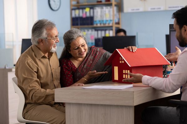 Elderly couple discussing a red house model with a professional at an office.