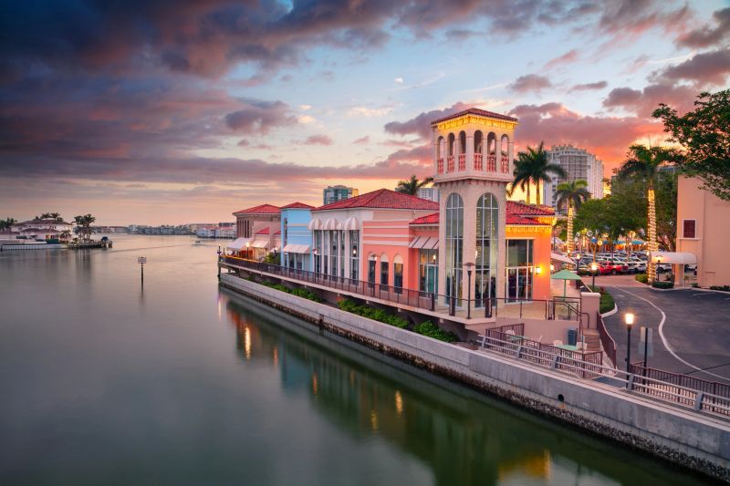 A scenic waterfront view showcases pastel-colored buildings with red-tiled roofs and a distinctive tower. Tall palm trees line the area, reflecting the tropical setting. The calm water beside the structures mirrors the warm hues of a stunning sunset, casting a soft glow over the scene. The sky is a mix of orange, pink, and purple tones, enhancing the tranquil atmosphere of Naples, Florida.