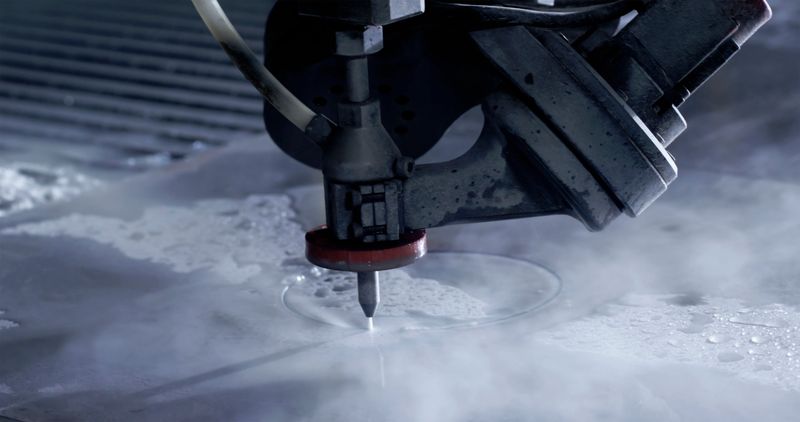 A close-up view of a water cutting CNC machine operating in a modern industrial setting. The machine precisely cuts through metal with high-pressure water jets under dim lighting conditions.