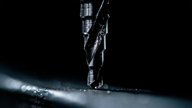 Macro photograph of industrial drill bit penetrating aluminum surface, creating fine metal shavings under dramatic spotlight against dark background. Sharp detail highlights precision metalworking process.