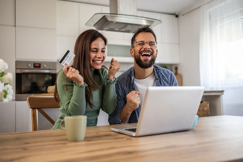 Young couple cheering while gazing at laptop, joyfully holding a credit card, celebrating the thrill of online shopping or the excitement of winning an auction from their cozy kitchen.