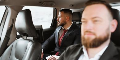 Two men in suits sitting inside a car, one in the back seat and the other in the front.
