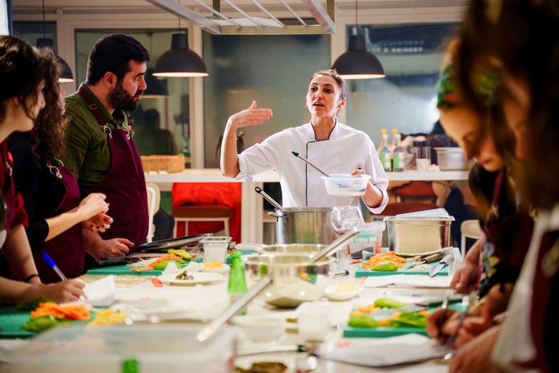 A professional female chef in a white uniform passionately instructs a diverse group of students in a hands-on cooking class. The participants, wearing aprons, attentively take notes and prepare ingredients on cutting boards. The well-lit, modern kitchen is filled with fresh vegetables, stainless steel cookware, and various cooking utensils, creating a dynamic and engaging learning environment.