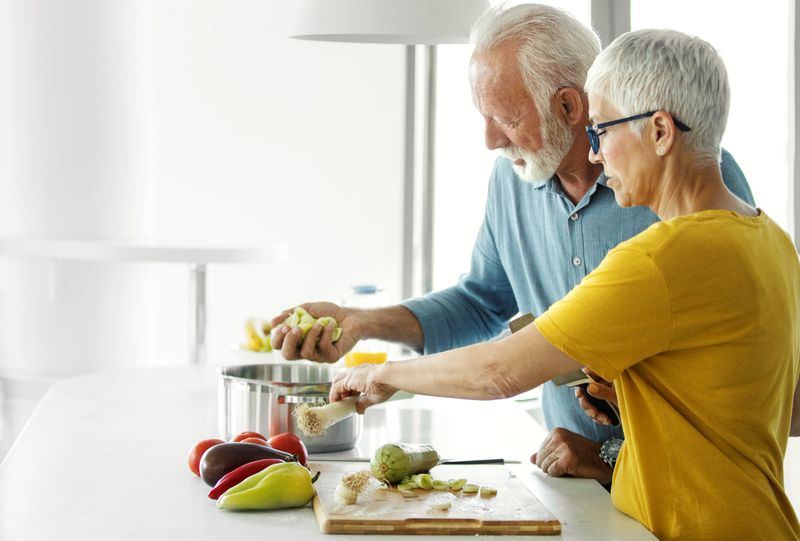 Closeup side view of a mid 60's couple having fun while preparing lunch in a brightly lit white colored kitchen. Bearded gentleman is stirring the soup while the lady is chopping vegetables.