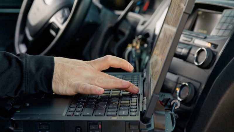 View of the interior of a police car, where a police officer sitting in the driving seat is using a laptop.