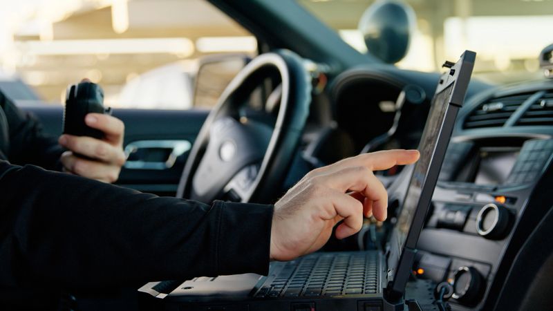 View of the interior of a police car, where a police officer sitting in the driving seat is using a laptop.