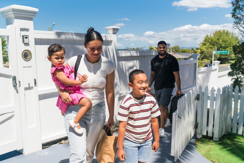 Pacific Island family entering a white picket fenced property on a sunny day, with a mother holding a child and a man carrying a grocery bag.