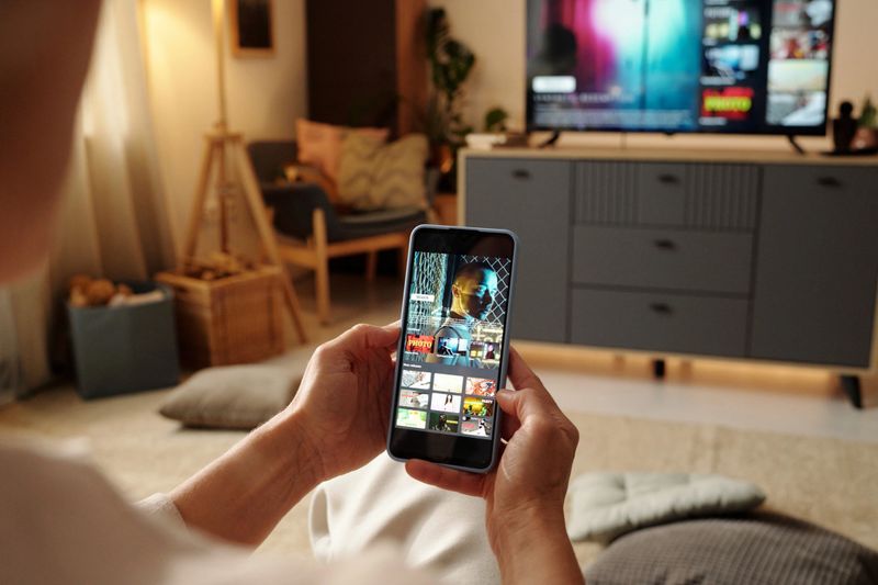 Hands holding smartphone while watching streaming videos in cozy living room with modern television set and comfortable furniture. Person enjoying multimedia content