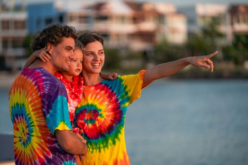 Three family members dressed in tie-dye shirts embrace each other by the water, looking out toward the colorful sunset while sharing laughter and smiles.