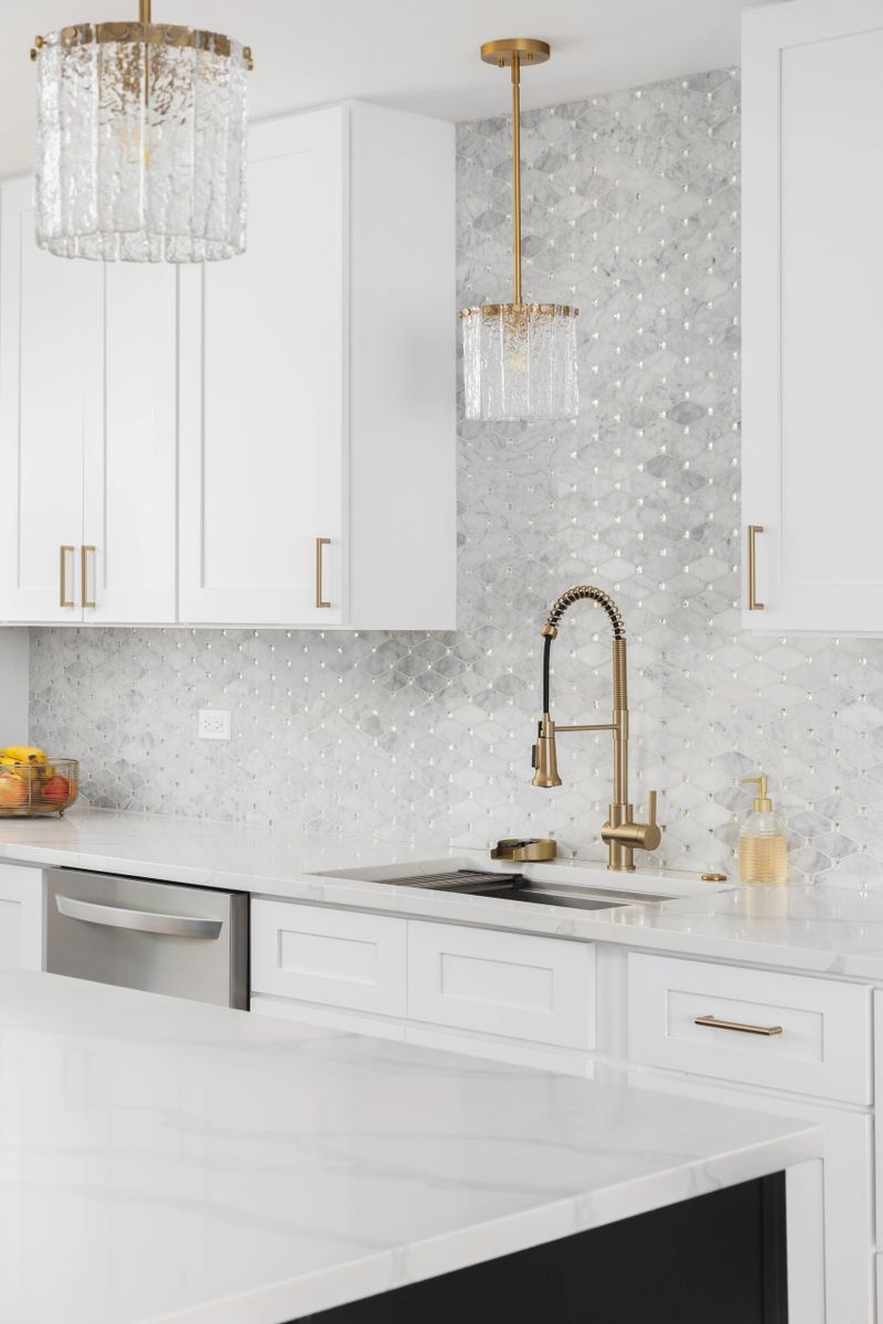 A kitchen with a gold faucet, white marble mosaic tile backsplash, white cabinets, and a white countertop.