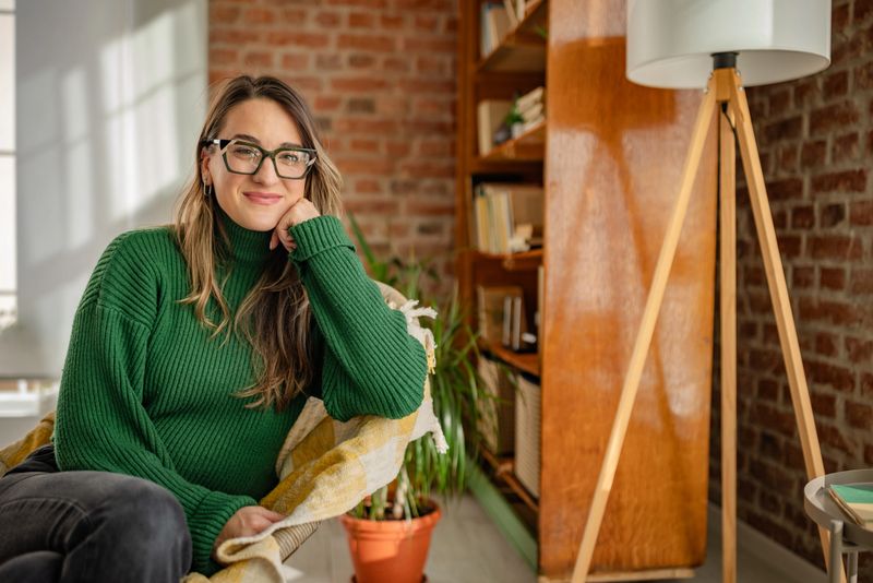 A young woman smiles while sitting comfortably at home in a modern living room. Surrounded by cozy decor and plants, she appears content in her leisure time, enjoying a peaceful moment.
