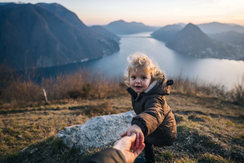 Personal perspective of toddler reaching for father's hand on rock above lake at sunset in winter setting, Ticino