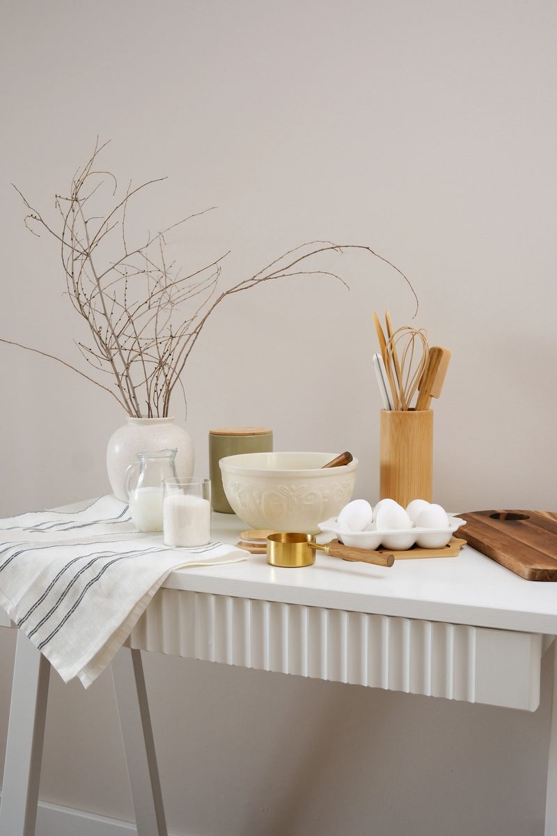 A curated collection of kitchen utensils, vase with dry branches and fresh ingredients: bowl, eggs, milk, sugar, cutboard, napkin is elegantly displayed on a neutral white table. High quality photo