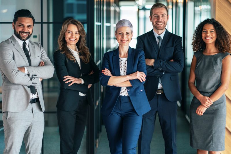 A group of five individuals dressed in formal business attire stands confidently in a contemporary office environment. They face the camera with serious and professional expressions, embodying teamwork and professionalism. The soft lighting enhances the atmosphere, while the blurred background suggests a sleek, modern workspace. This image captures the essence of corporate collaboration and the dedication of professionals in a dynamic business setting.