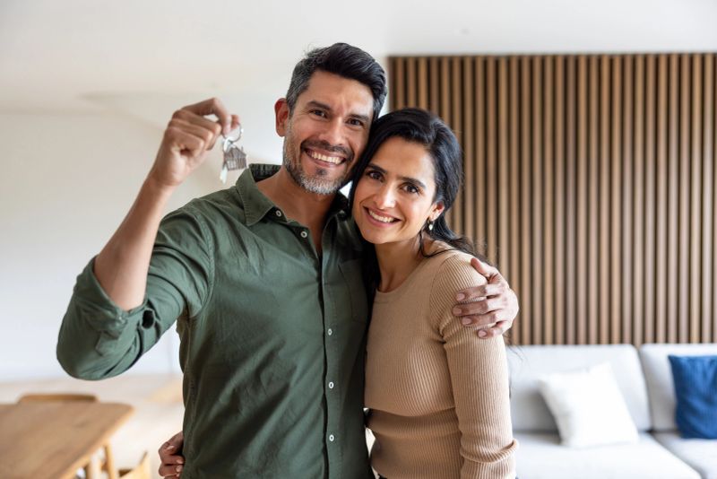 Loving Latin American couple smiling while holding the keys of their new house - home ownership concepts