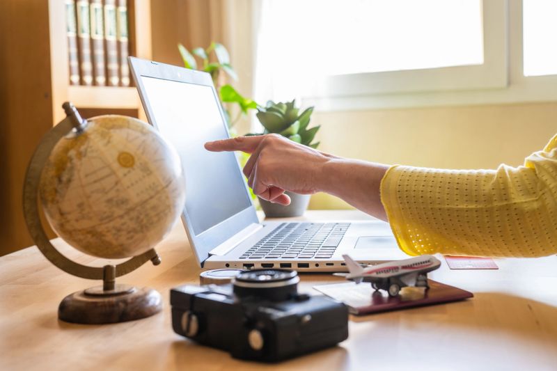 Woman pointing at laptop screen while planning a trip on a booking website, surrounded by a globe, camera, passport, and airplane model on the desk, creating an organized itinerary