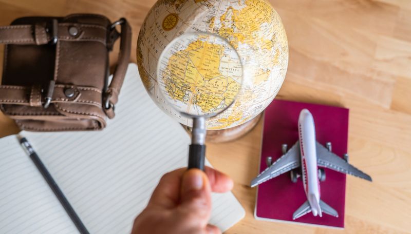 Traveler planning next trip using magnifying glass on globe, passport, airplane, and notebook, organizing world tour, travel schedule, and holiday concept