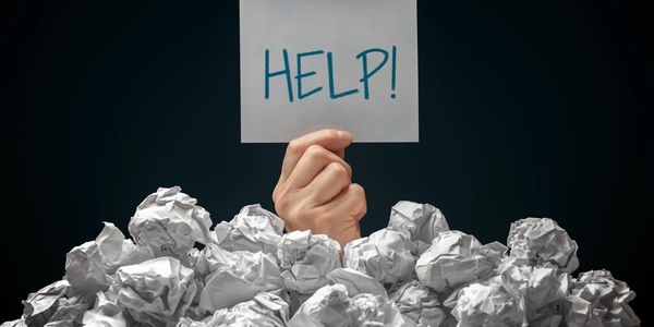 Hand holding a HELP sign above crumpled paper pile.