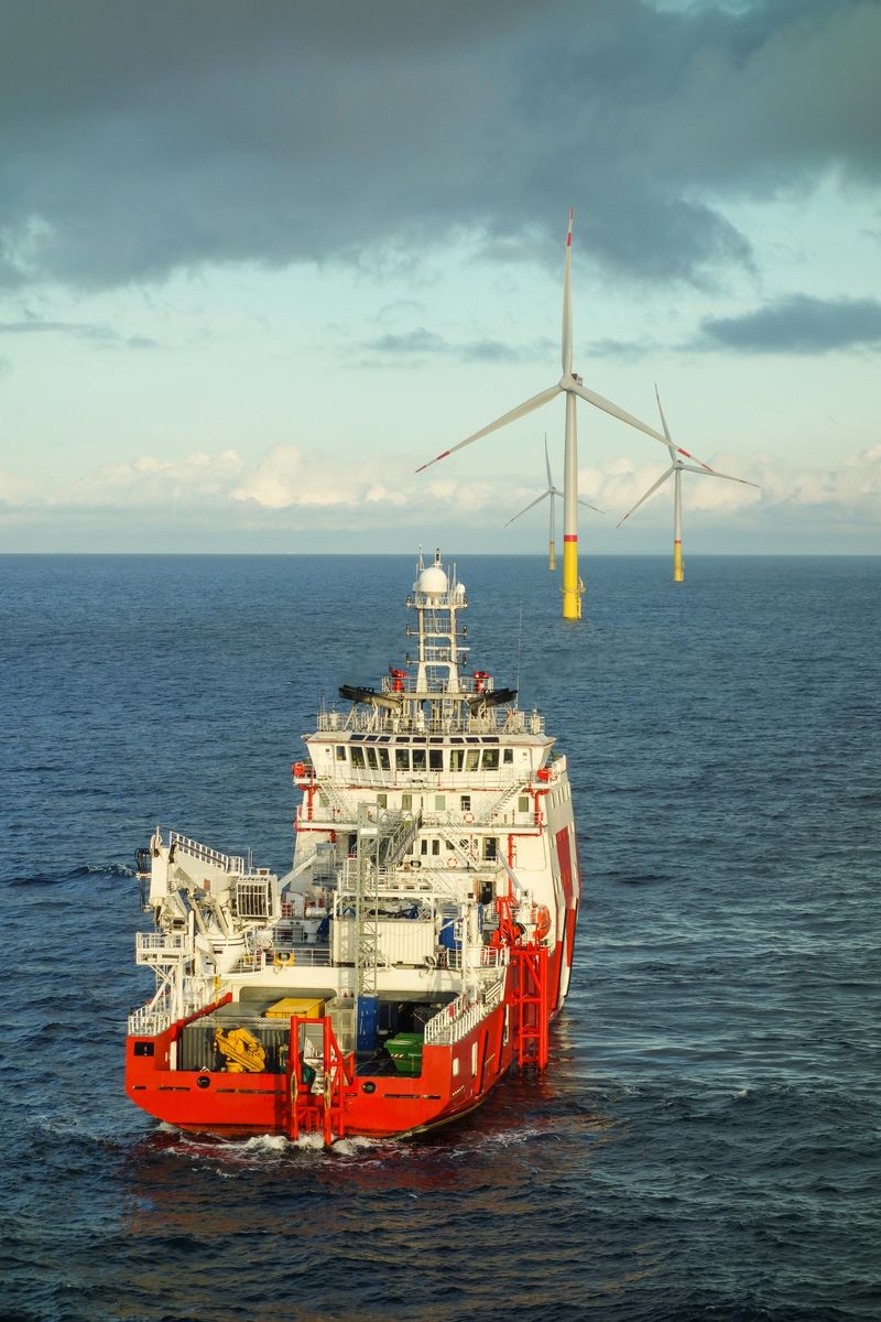 A stunning view of an offshore wind farm in Germany, where massive wind turbines rise above the open sea. Large service vessels navigate front of the turbines. The scene captures the scale and complexity of offshore renewable energy operations, with dramatic lighting and ocean conditions adding to the epic atmosphere.